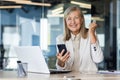 Portrait of a senior gray-haired businesswoman working in the office at a laptop, holding a phone in her hands, happy Royalty Free Stock Photo
