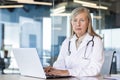 Portrait of a senior gray-haired female doctor sitting in the office, working online on a laptop and looking seriously Royalty Free Stock Photo