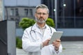 Portrait of a senior gray-haired male doctor in a white coat and with a stethoscope standing outside the clinic, holding Royalty Free Stock Photo