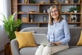 Portrait of senior gray-haired woman at home. A woman is sitting on a sofa in the living room, smiling and looking at Royalty Free Stock Photo