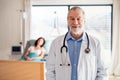 Portrait of senior male doctor standing in hospital room, patient in the background. Royalty Free Stock Photo