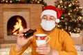 Portrait of senior man wearing sweater and santa claus hat, holding mug and syrup, sitting in festive living room near fireplace Royalty Free Stock Photo