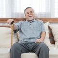 Portrait of senior old elderly asian man sit on coach hand hold help walking stick sit on sofa in house look at camera with