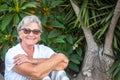 Portrait of senior pretty woman with gray hair. Sitting outdoor in the garden. Background of plants and leafs. Smiling and