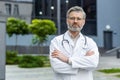 Portrait of senior serious doctor outside modern clinic outdoors, mature man in medical coat looking at camera thinking Royalty Free Stock Photo