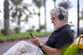 Portrait of a senior white-haired man with beard sitting in a bench and listening to music with headphones. Handsome people using Royalty Free Stock Photo