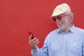 Portrait of senior white haired man with cap smiling using his smart phone, standing against red background Royalty Free Stock Photo