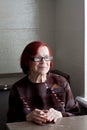 Portrait of senior woman with red hair, wearing glasses, in kitchen, sitting at table. elderly woman looks out window and smiles.