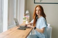 Portrait of serious attractive young female freelancer typing text on keyboard using laptop sitting at table by window Royalty Free Stock Photo