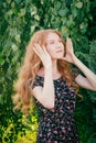 Portrait of a serious beautiful redhead lady in summer dress fixing her hair with hands under birch tree in spring time Royalty Free Stock Photo