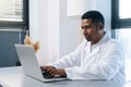 Portrait of serious black male doctor wearing white coat uniform using working on laptop computer sitting at desk in Royalty Free Stock Photo