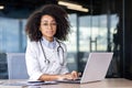 Portrait of a serious confident female doctor sitting in a hospital office at a desk, working on a laptop and looking at Royalty Free Stock Photo