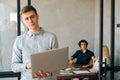 Portrait of serious confident young businessman in casual clothes holding laptop in hand standing at office looking at Royalty Free Stock Photo