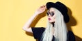 Portrait of serious fashion hipster blonde girl, wearing sunglasses, touching her hat, isolated on yellow background. Royalty Free Stock Photo