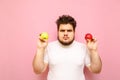 Portrait of a serious fat man with red and green apple in his hands stands on a pink background, looks in camera with a serious Royalty Free Stock Photo