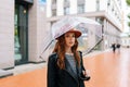 Portrait of serious ginger young woman wearing elegant hat standing with transparent umbrella on street in rainy weather Royalty Free Stock Photo