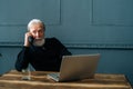 Portrait of serious gray-haired senior adult business man talking on mobile phone sitting at wooden table with laptop Royalty Free Stock Photo