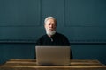 Portrait of serious gray-haired senior aged male sitting at wooden table at home office working using computer laptop Royalty Free Stock Photo