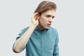 Portrait of serious handsome long haired blonde young man in blue casual shirt standing, holding hand on ear and trying to hear or Royalty Free Stock Photo