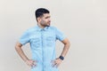 Portrait of serious handsome young bearded man in blue shirt standing, with hands on waist and looking away with thoughtful face Royalty Free Stock Photo
