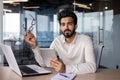 Portrait of a serious Indian young man businessman sitting in the office at the desk and looking at the camera, holding Royalty Free Stock Photo