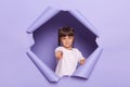 Portrait of serious little girl with pigtails wearing striped shirt breaks through purple paper background, female kid pointing