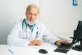 Portrait of serious mature adult male doctor in white coat with stethoscope working on desktop computer at desk in Royalty Free Stock Photo