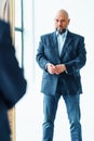 Portrait of serious middle-aged bald man with gray beard wearing blue suit jacket, looking gravely at mirror reflection.