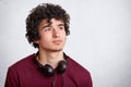 Portrait of serious pensive curly male teenager with curly hair, uses modern headphones for listening favourite music, looks pensi