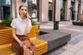 Portrait of serious pretty young woman having smartphone conversation sitting on wooden yellow bench on city street in Royalty Free Stock Photo