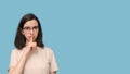 Portrait of a serious student girl with glasses, covering her mouth with her finger on a blue background. Royalty Free Stock Photo