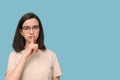 Portrait of a serious student girl with glasses, covering her mouth with her finger on a blue background. Royalty Free Stock Photo