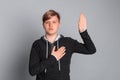 Portrait of serious teenager boy with hand on chest giving oath, promising to be honest and to tell truth, standing over grey