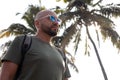Portrait of serious young bald bearded handsome man wearing khaki T-shirt, blue sunglasses, rucksack, standing outdoor. Royalty Free Stock Photo