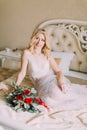 Portrait of blonde bride in white underwear sitting on bed. Bouquet with red roses Royalty Free Stock Photo