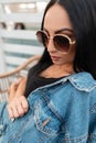 Portrait of a sexy pretty young woman with beautiful black hair in trendy sunglasses in a black t-shirt in a stylish denim jacket Royalty Free Stock Photo