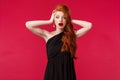 Portrait of shocked and ambushed young redhead female in elegant black dress, gasping nervously, look alarmed camera