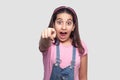 Portrait of shocked beautiful brunette young girl in casual pink t-shirt and blue overalls standing, looking and pointing at camer