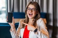 Portrait of shocked beautiful stylish young woman in glasses sitting, showing and pointing at mobile smart phone screen and Royalty Free Stock Photo