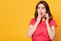 Portrait of shocked emotional girl holding mobile phone in one hand, covering opened mouth with hand, looking aside, wearing Royalty Free Stock Photo