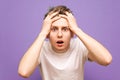 Portrait of a shocked guy on a purple background. Young man in a white shirt took his hands behind his head and emotionally looks