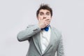 Portrait of shocked handsome bearded man in casual grey suit and blue bow tie standing covering his mouth and looking at camera Royalty Free Stock Photo