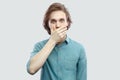 Portrait of shocked handsome long haired blonde young man in blue casual shirt standing, covering his mouth and looking at camera Royalty Free Stock Photo