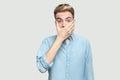 Portrait of shocked handsome young man in light blue shirt standing with scared face, covering his mouth and looking at camera Royalty Free Stock Photo