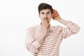 Portrait of shocked intrigued attractive caucasian man in trendy striped shirt, holding hand near ear and mouth Royalty Free Stock Photo