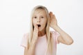 Portrait of shocked intrigued chatty young girl with long blond hair, holding hand near ear and opened mouth, saying wow