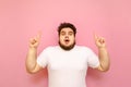 Portrait of shocked overweight man on pink background, looks into camera with surprised face and shows hands up on copy space. Royalty Free Stock Photo