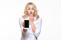 Portrait of shocked pretty blond business woman with hand on her mouth showing mobile phone blank screen isolated over white.