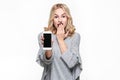 Portrait of shocked pretty blond woman with hand on her mouth showing mobile phone blank screen isolated over white background.