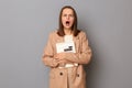 Portrait of shocked scared woman wearing beige jacket standing with organizer and calculator in hands isolated over gray Royalty Free Stock Photo